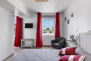 A bedroom with a bed, a chair, a small table, a TV, and a window with curtains.