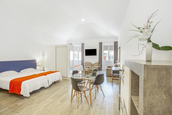 Ein Hotelzimmer mit einem Bett, einem Esstisch mit Stühlen, einem Fernseher und einer Vase mit Blumen.