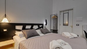 A neatly made bed with pillows, a bedside table, a hanging light fixture, a mirror, and a door with a towel rack.