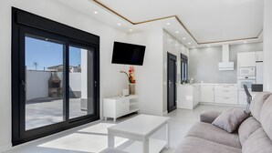 Una sala moderna con un sofá, una mesa de centro, un televisor y una cocina con armarios y electrodomésticos blancos.
