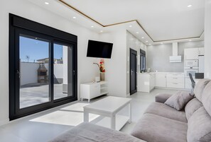 Ein modernes Wohnzimmer mit einer Couch, einem Couchtisch, einem Fernseher und einer Küchenzeile mit weißen Schränken und Geräten.