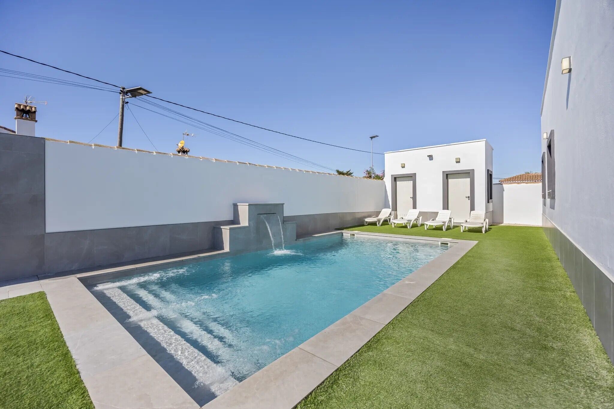 Une piscine extérieure moderne, agrémentée d’une petite cascade, entourée d’une pelouse bien entretenue et d’un bâtiment blanc doté d’une porte et de fenêtres.
