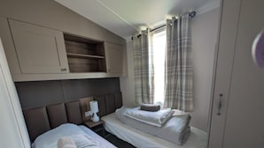 A bedroom with two beds, a bedside table with a lamp, a wardrobe, and a window with curtains. - Seaside Hunstanton Manor Park 2026 Model (Hunstanton)