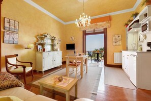 Een woonkamer met een bank, stoelen, een salontafel, een kroonluchter, een eethoek en een keuken met witte kastjes.
