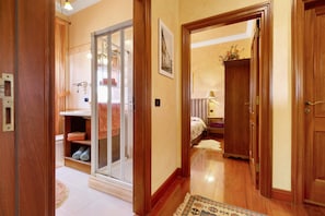 A hallway with wooden doors, a glass door leading to a bathroom, and a bed in the background.