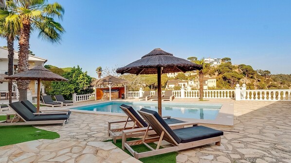 Uma área de piscina com espreguiçadeiras e guarda-sóis, cercada por piso de pedra e vegetação.
