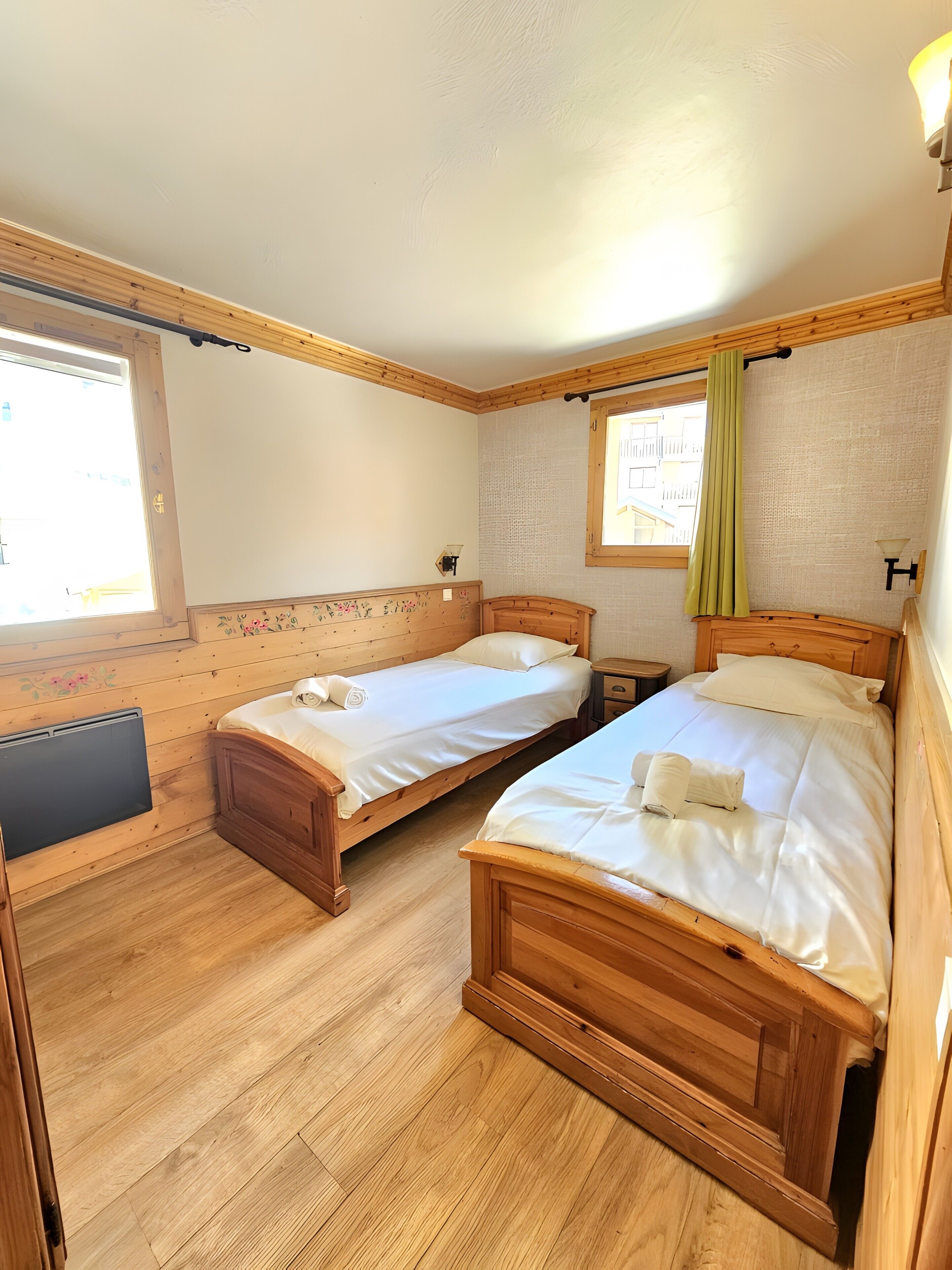 A room with two wooden beds, a small table, a radiator, and a window with a curtain.