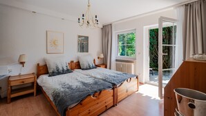 A bedroom with a wooden bed, a nightstand, a lamp, a chandelier, a window with a view of greenery, and a door leading outside. - Apartment 'Rai Mattiss' with Mountain View, Private Terrace and Wi-Fi (Oberharmersbach)