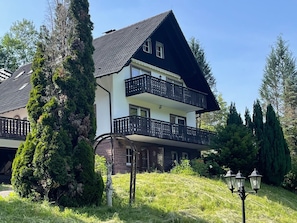 A two-story house with a balcony, surrounded by greenery and a lamp post.
