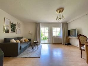 A living room with a sofa, a coffee table, a TV on a stand, a chandelier, and a view through the windows.