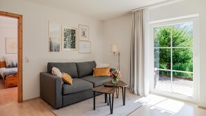 A modern living room with a grey sofa, a small coffee table, and a floor lamp. There are framed pictures on the wall and a large window with a view of greenery. - Apartment 'Rai Mattiss' with Mountain View, Private Terrace and Wi-Fi (Oberharmersbach)