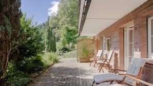 A stone pathway leading to a seating area with wooden chairs and a white cushioned bench, surrounded by greenery and a building with brick walls and large windows. - Apartment 'Rai Mattiss' with Mountain View, Private Terrace and Wi-Fi (Oberharmersbach)