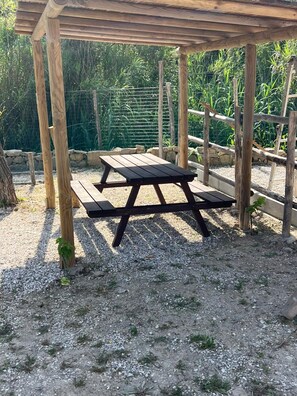 Uma pérgola de madeira com uma mesa de piquenique por baixo, rodeada por cascalho e vegetação.
