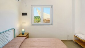 A bedroom with a bed, a bedside table with a lamp, a window with a view, and a wooden shelf with towels. - Calipso Apartment with Private Garden and Air Conditioning (Casal Velino)