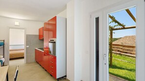 A compact kitchen with red cabinets, a dining area with a table and chairs, and a sliding glass door leading to a garden. - Calipso Apartment with Private Garden and Air Conditioning (Casal Velino)