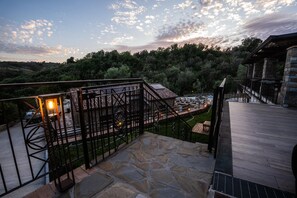 Um terraço de pedra com guarda-corpo metálico, vista para uma área arborizada e um edifício com telhado inclinado.