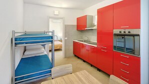 A compact kitchen with red cabinets, a sink, and a built-in oven. There is a bunk bed with blue bedding in the adjacent room. - Calipso Apartment with Private Garden and Air Conditioning (Casal Velino)