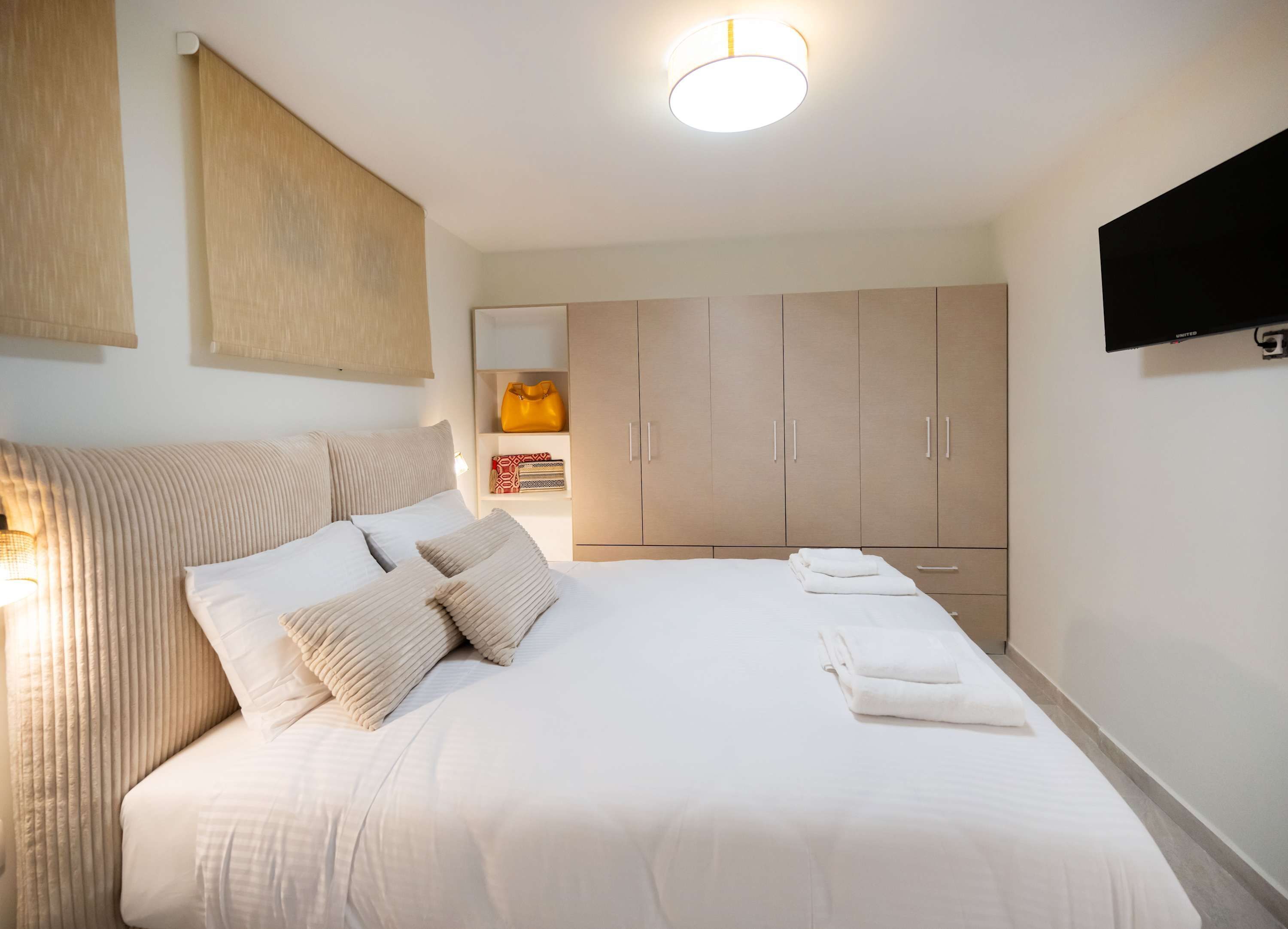 A neatly arranged bedroom with a bed, bedside lamps, a wardrobe, and a flat-screen TV mounted on the wall.