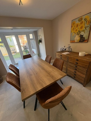 A dining area with a wooden table, cushioned chairs, a cabinet with drawers, a painting of sunflowers, and a bicycle. - Spacious Modern Home in Whitefield (Bury)