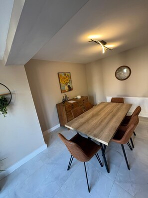 A dining area with a wooden table, brown chairs, a clock, and a painting on the wall. - Spacious Modern Home in Whitefield (Bury)