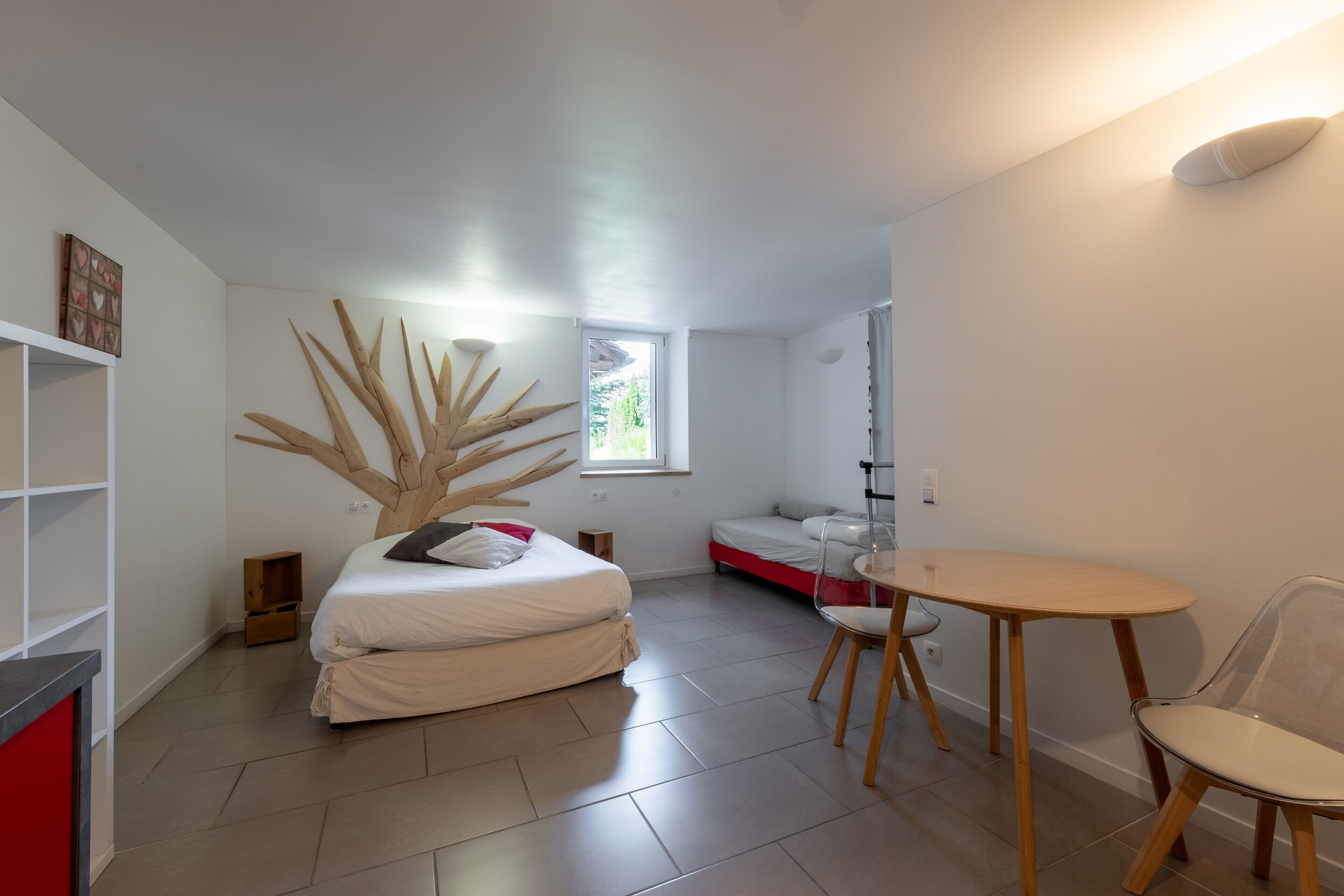 Une chambre moderne avec un lit, une petite table et une chaise. Il y a une fenêtre donnant sur des arbres.