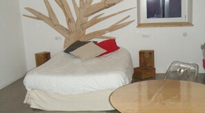 A bed with a wooden tree-like structure on top, a white pillow, a red pillow, a small wooden table, and a chair.