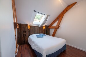 A bedroom with a sloped ceiling, a skylight, wooden beams, a bed with white bedding, and a small wooden shelf. - Gîte l'Evasion, Group Accommodation for 20 to 35 Guests (Bussang)