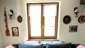 A living room with a sofa, pillows, a round table, a window with curtains, wall decorations, and a tiled floor. - Holiday Home “La Maison De Mediovilla” with Private Terrace, Garden & Wi-Fi (Wingersheim les Quatre Bans)