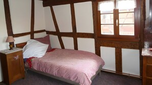 A bedroom with exposed wooden beams, a bed with a checkered bedspread, a nightstand with a lamp, and a window with a view of buildings. - Holiday Home “La Maison De Mediovilla” with Private Terrace, Garden & Wi-Fi (Wingersheim les Quatre Bans)