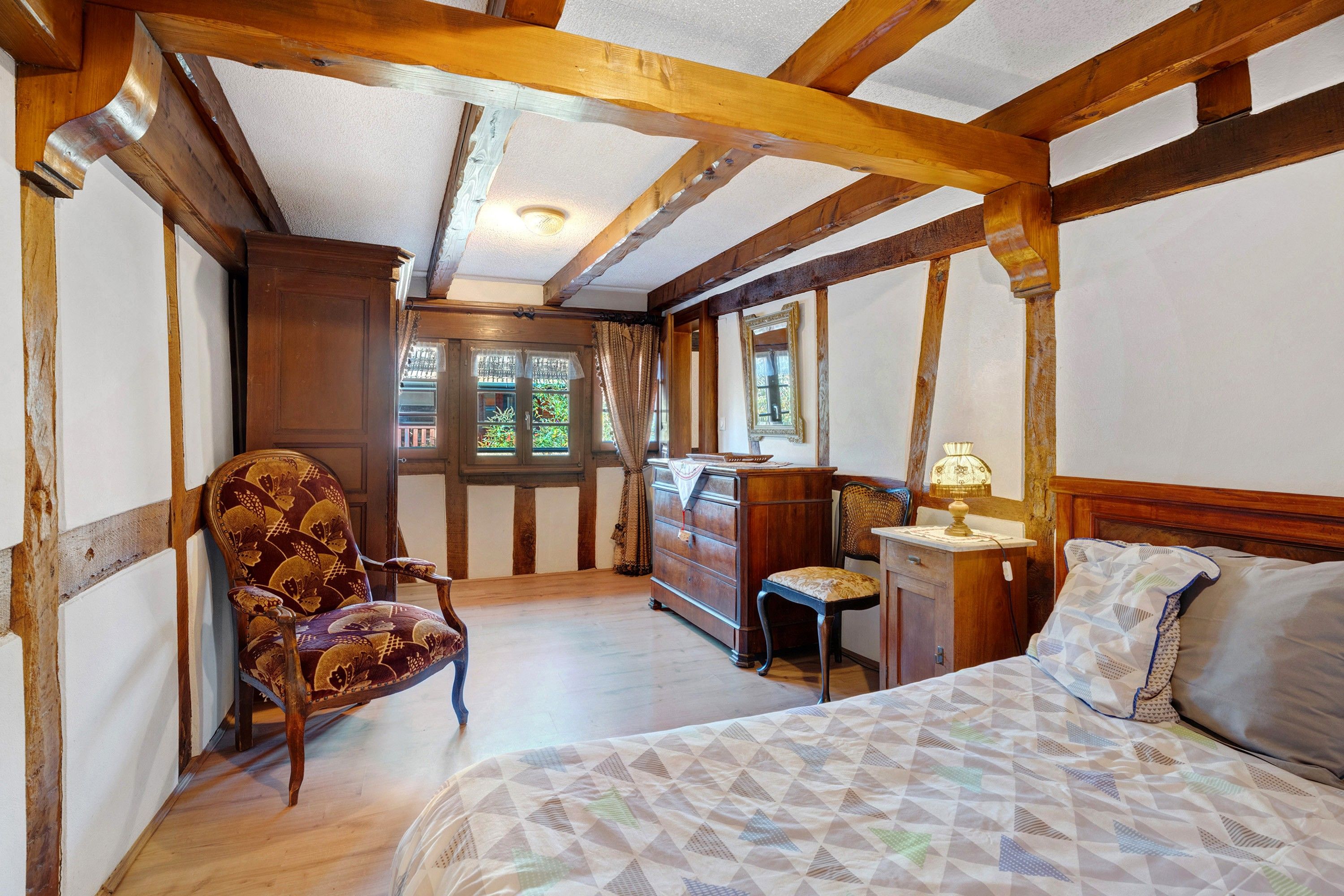 Ein Zimmer mit freiliegenden Holzbalken, einem Bett mit gemustertem Überwurf, einem Sessel, einem Schminktisch und einem Fenster mit Vorhängen.