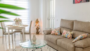 Un salon avec un canapé beige, des coussins colorés, une table basse en verre, une table à manger avec des chaises et une statue décorative.