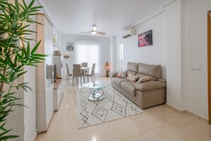 Uma sala de estar com sofá, mesa de centro de vidro, área de jantar com cadeiras, ventilador de teto e ar-condicionado fixado na parede.