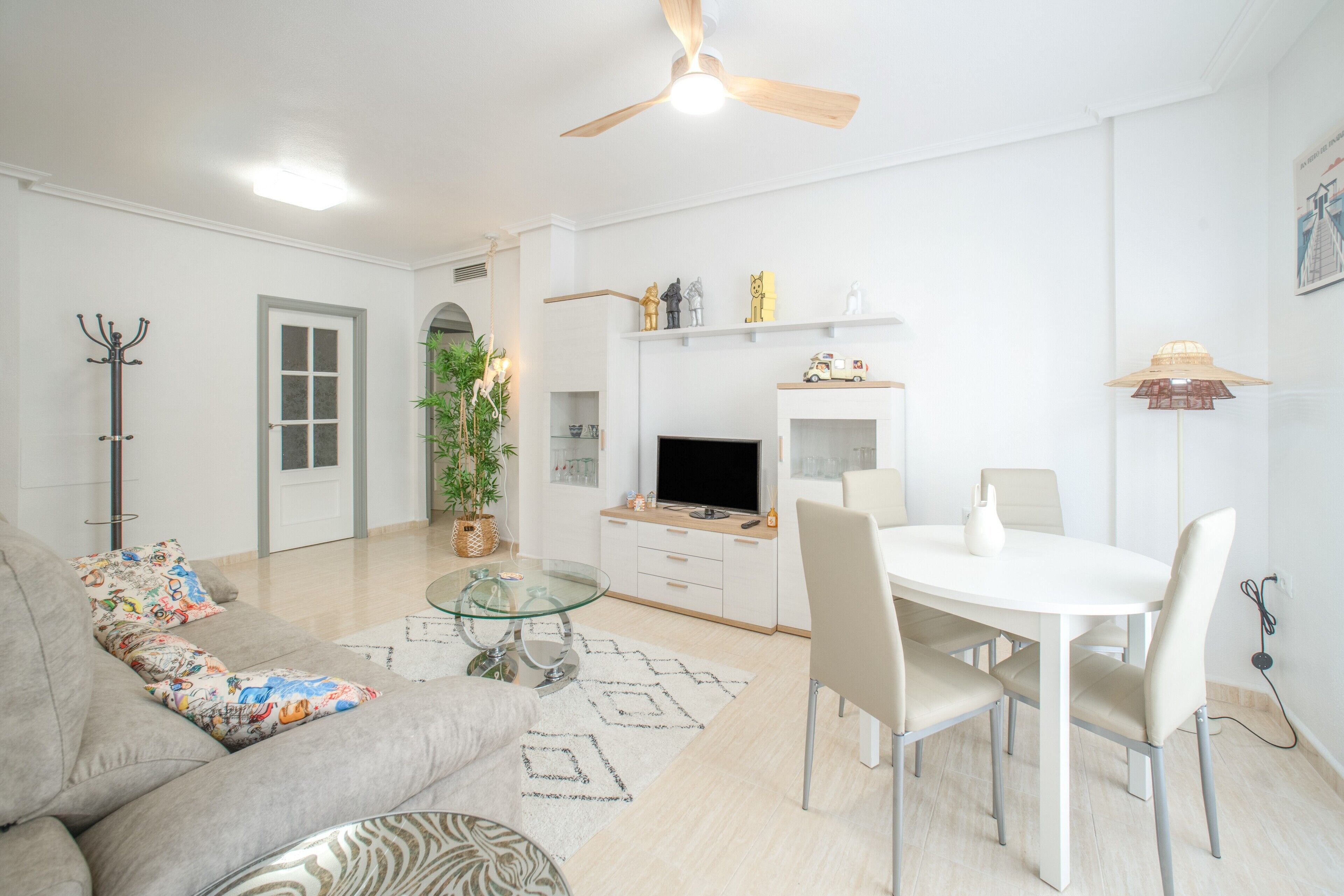 Ein modernes Wohnzimmer mit Glastisch, einer Couch mit bunten Kissen, einem Essbereich mit weißem Tisch und Stühlen, einem Fernseher und einem Deckenventilator.