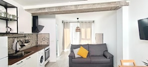 Ein kompakter Wohnbereich mit Küchenzeile, einem grauen Sofa und einem großen Fenster.