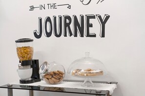 A breakfast setup with a cereal dispenser, a bowl of cookies, and a covered dessert display on a table against a wall with the text 'IN THE JOURNEY'. - Camera Mare - Casa Del Capitano, with Wi-Fi and Air Conditioning (Chioggia)