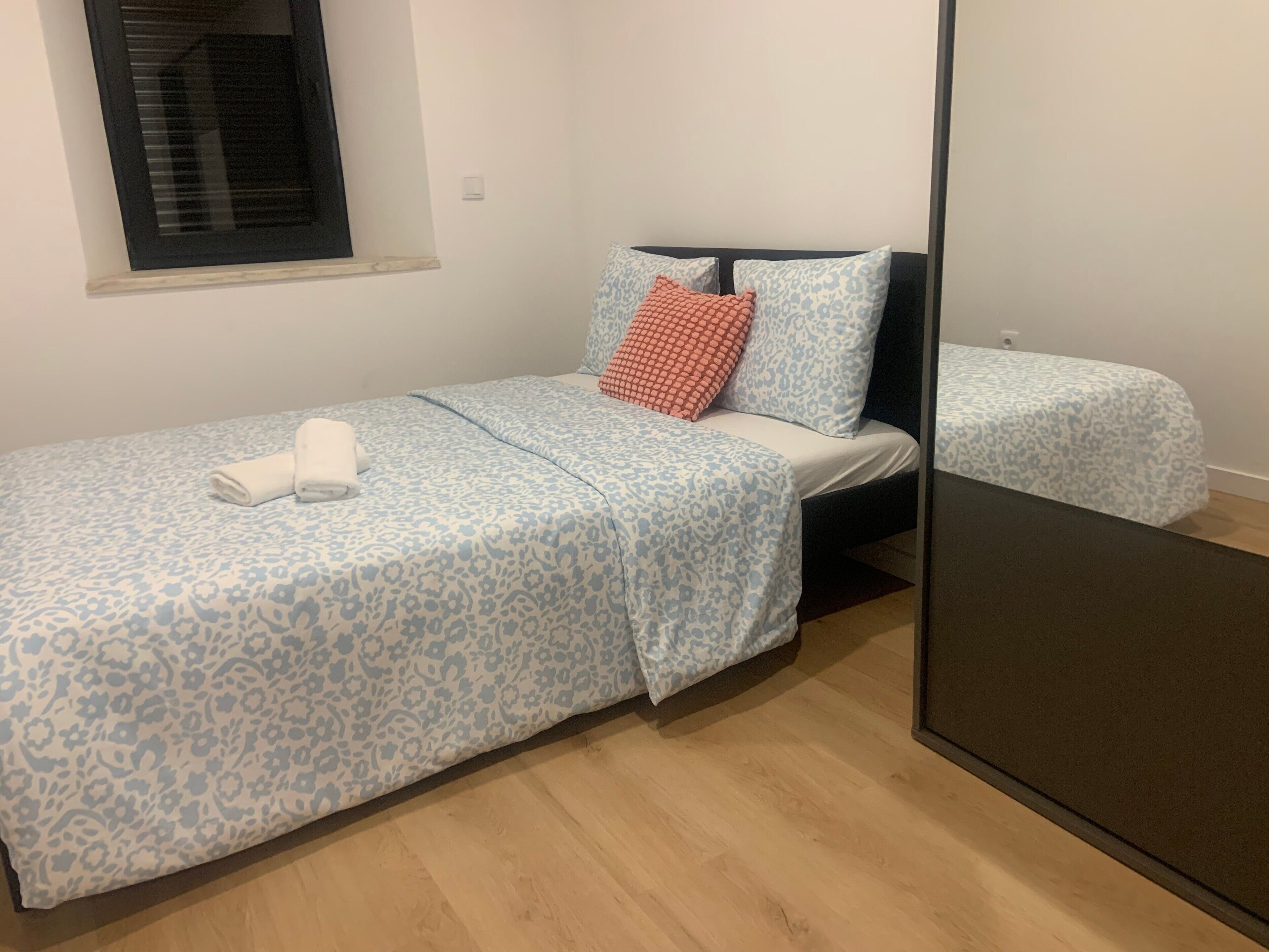 A bedroom with a bed, a window, a mirror, and a wooden floor.