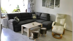 Ein modernes Wohnzimmer mit einer Eckcouch, einem kleinen Tisch, zwei Hocker, einem Sessel und Kunstwerken an der Wand.
