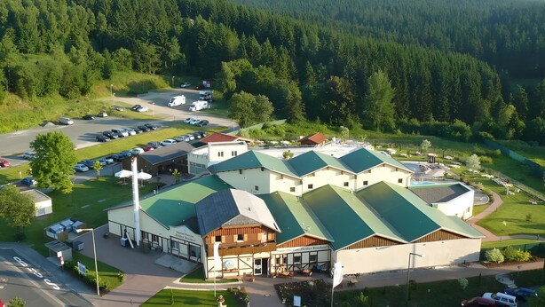 Ein Hotel mit Gründach, Parkplatz und bewaldeter Landschaft im Hintergrund.