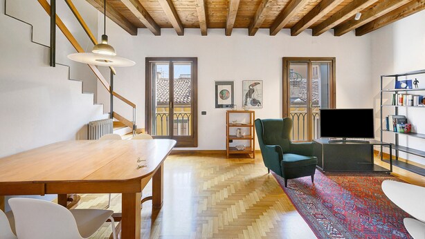 Een ruime woonkamer met houten plafond, een eettafel, stoelen, een televisie en een boekenkast.