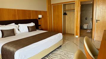 A hotel room with a large bed, a wooden headboard, a bedside table, a lamp, a wardrobe, and a bathroom visible through an open door.