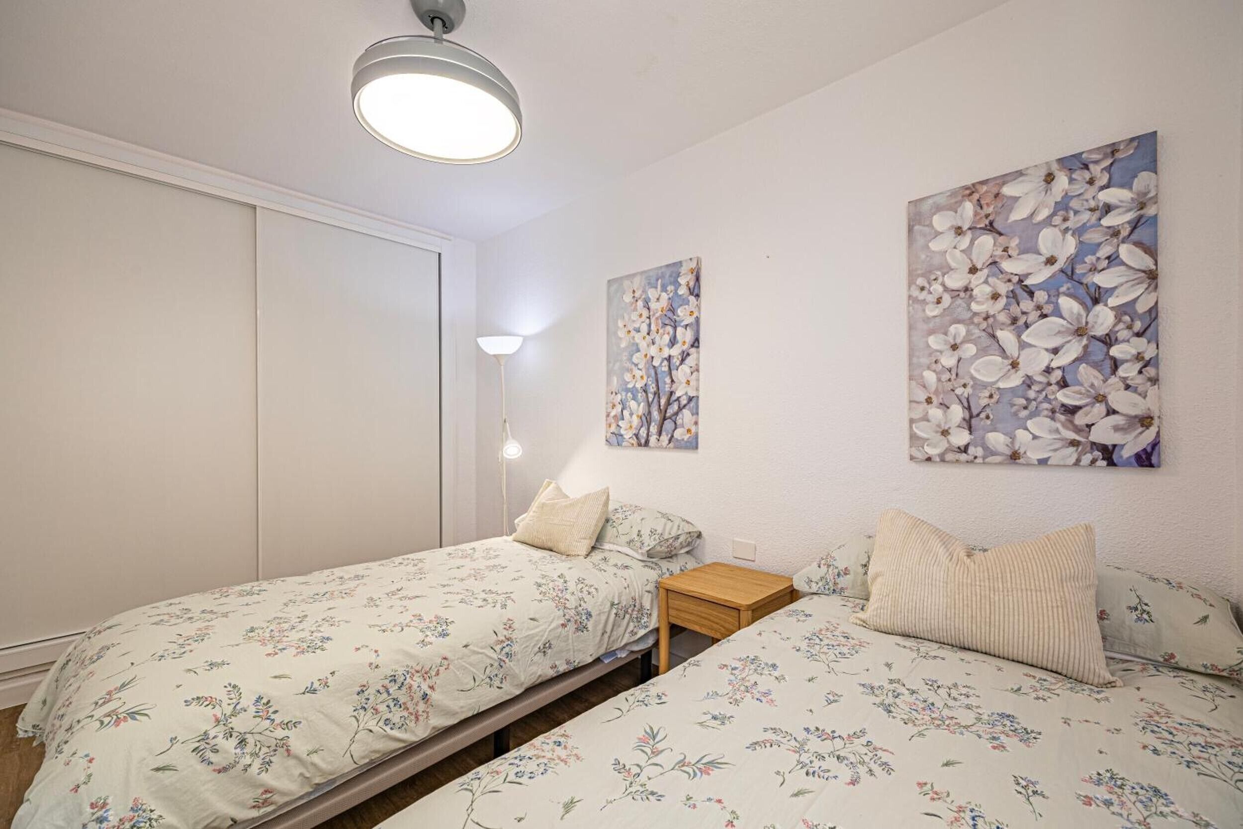 A bedroom with two beds, a bedside table, a lamp, and floral artwork on the wall.