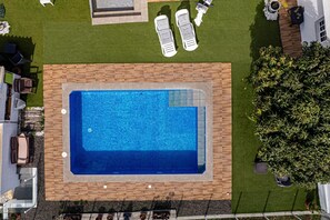 Une vue aérienne d’une piscine entourée d’une terrasse en bois, avec des chaises longues et un petit espace aménagé en jardin.