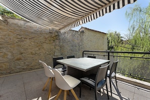 An outdoor patio with a table and chairs, a striped awning, stone walls, and a view of greenery.