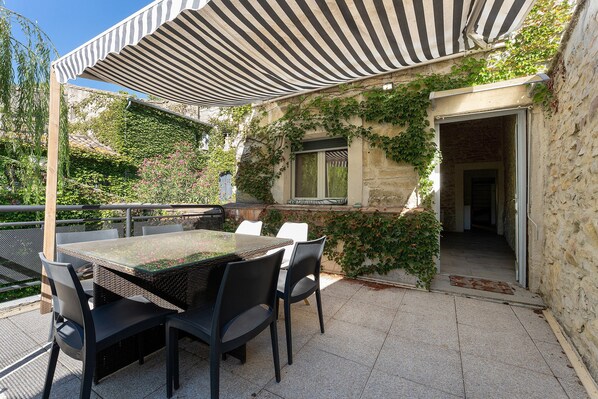 An outdoor patio with a table and chairs, a striped awning, and a stone wall with greenery.