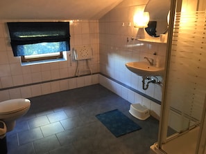 Et lille badeværelse med toilet, vask, brusebad og et vindue med gardin.