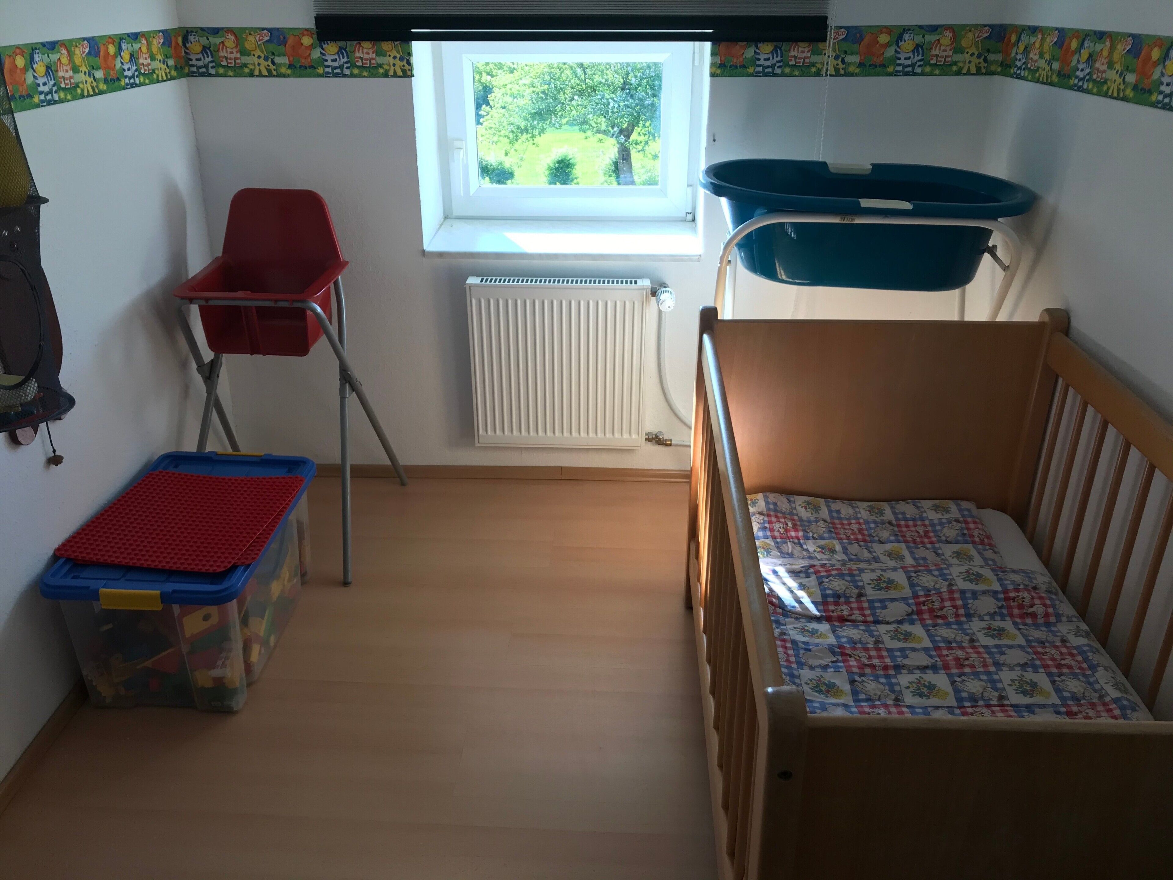 Ein Kinderzimmer mit einem hölzernen Bett, einem roten Stuhl, einer Aufbewahrungsbox, einem Fenster mit Blick ins Grüne und einer Heizung.