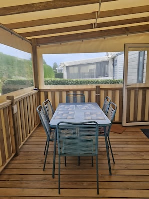 Zona exterior coberta amb taula i cadires, terrassa de fusta i vista a una casa i vegetació.