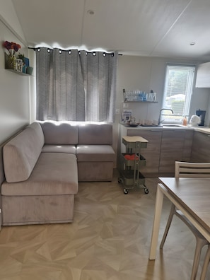 Ein modernes Wohnzimmer mit einer Eckcouch, einem Esstisch, einer Küchenzeile inklusive Spüle und einem Fenster mit Jalousien.