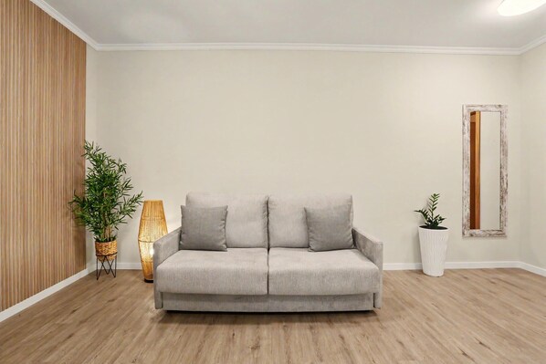 A neutral-toned sofa with cushions, a wicker floor lamp, and a potted plant in a corner of a room with wooden flooring and a plain wall. - Apartment 'Ninho Ilhéu' with Mountain View and Wi-Fi (Câmara de Lobos)