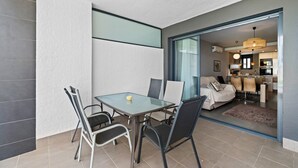 Un salon moderne avec une table et des chaises en verre, un canapé et une cuisine visible à travers une porte ouverte.